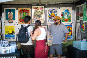 Three people stand at an outdoor art booth, talking. Colorful tattoo-style prints hang behind them. Two visitors with backpacks face an artist wearing a hat and sunglasses. Stickers and art are on tables.
