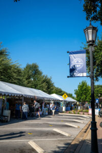 Outdoor market with white tents and shoppers under a clear blue sky. A streetlamp and blue Downtown banner are visible, with trees lining the street and a sidewalk on the right.