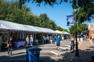 People browse art booths under white tents at an outdoor street fair on a sunny day. A blue city banner hangs from a lamp post, and trees line the background. A blue recycling bin is visible in the foreground.