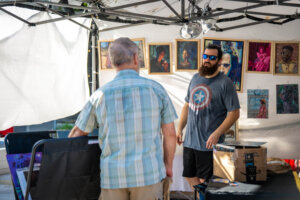 A man wearing a Captain America shirt and sunglasses stands at an art booth, talking to another man. Portrait paintings are displayed on the wall behind them.
