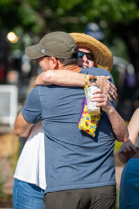Two people embrace outdoors on a sunny day. One wears a straw hat and sunglasses, holding a drink with a colorful holder, while the other wears a cap and blue shirt. Trees are visible in the blurred background.
