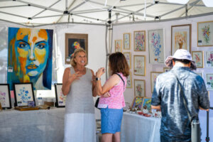 Two women talk in an outdoor art booth displaying colorful paintings and framed floral artwork, while a man with a hat browses nearby.