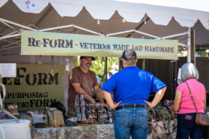 A vendor smiles while talking to two people at an outdoor booth with a sign reading Re-Form - Veteran Made and Handmade, displaying handmade items under a white tent.