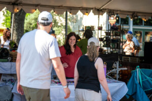 A woman stands behind a vendor table at an outdoor market, smiling and talking to two people in casual clothes and hats who approach her booth under a white tent.