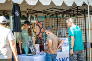 A woman in a green shirt and cap smiles at two people at an outdoor booth under a white tent. The table displays flyers and a t-shirt hangs nearby. The setting appears to be a community event or festival.