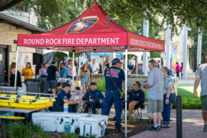 Several people gather under a red Round Rock Fire Department tent at an outdoor event. Firefighters sit and stand, talking with attendees. Coolers, gear, and a yellow kayak are visible. Trees provide shade.