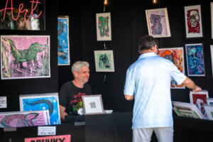 An artist sits at a booth displaying colorful animal prints, while a man in a light shirt browses through a bin of artwork. The booths backdrop is black, highlighting the vibrant framed prints.
