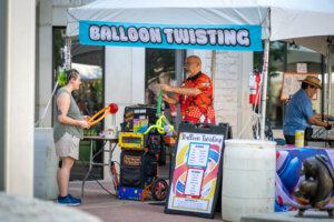 A person makes balloon animals for a woman under a Balloon Twisting tent at an outdoor event; colorful balloons and a price list are visible in front.