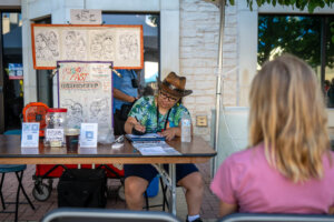 A caricature artist wearing a hat draws a seated customer at an outdoor booth. Caricature samples hang on display, and a sign advertises $5 per drawing. Various items and tip jars are on the table.