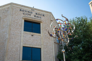 Round Rock City Hall building with tan stone facade, blue windows, and address number 221. In front is a modern metal and colored glass sculpture against a clear blue sky.