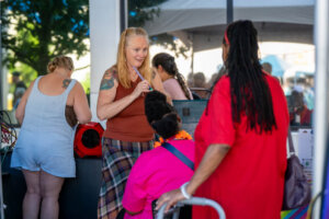 A woman with long red hair and a plaid skirt smiles and talks to two women, one seated, at an outdoor event booth. Another person stands nearby, facing away. Trees and a tent are visible in the background.