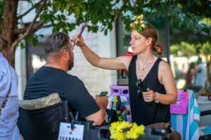 A woman with a floral headpiece paints a mans face outdoors. She holds a brush while the man sits still, holding his phone. There are art supplies and colorful decorations on the table beside them.