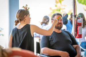 A woman paints a rainbow on a smiling mans face at an outdoor event. Other people are visible in the background, and a fire extinguisher hangs nearby.
