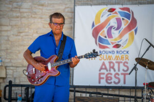 A man in a blue outfit plays an electric guitar on stage at the Round Rock Summer Arts Fest, with a logo banner and drum set in the background.