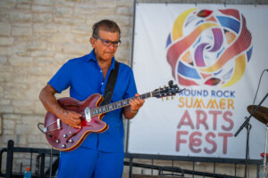 A man in a blue outfit plays an electric guitar onstage beside a sign that reads Round Rock Summer Arts Fest. A brick wall is in the background.