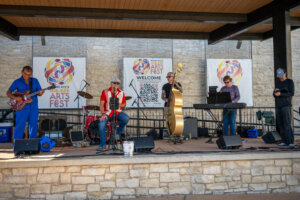 A five-member band performs on an outdoor stage with instruments, including guitar, drums, keyboard, and upright bass, at a summer arts festival. Festival banners and a QR code are displayed on the stone wall behind them.