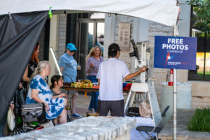 A group of people, including a woman in a wheelchair, pose for a photo at an outdoor booth with a Free Photos sign. A photographer stands behind a camera, while others wait nearby.