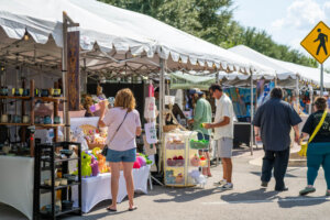 People browse booths with crafts and handmade items at an outdoor art market under white tents on a sunny day. A yellow pedestrian crossing sign is visible in the background.