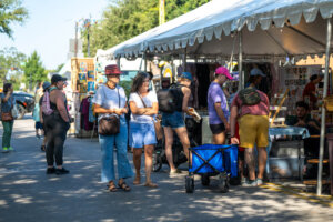 People browse booths and artwork at an outdoor market under a white tent on a sunny day. Some look at crafts, while others stand or walk along the pathway. Trees and displays are visible in the background.
