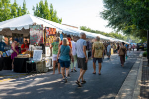 People walk along an outdoor art market with white tents and vendors selling artwork and crafts on a sunny day. Trees line the street, and shoppers browse various booths.