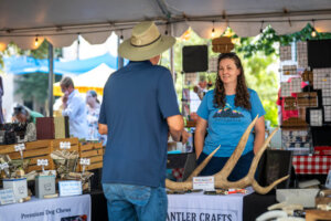 A woman stands behind a booth selling antler crafts and dog chews at an outdoor market, smiling and talking with a man wearing a hat. Tables display products and signs under a white tent.