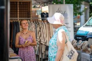 A woman in a purple dress smiles while standing at a clothing market stall, talking to another woman in a sun hat and aqua dress. Clothing racks and jewelry displays are visible around them.