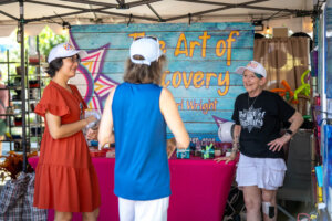 Three women wearing hats stand and chat at a booth with a bright pink table and a blue sign reading The Art of Recovery at an outdoor event. One woman is holding a plastic bag, and they all appear to be smiling.