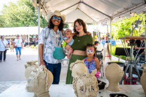 A family with two children stands under a tent at an outdoor event, posing by a table with clay sculptures. One child has face paint, and the parents are smiling. Other people and booths are visible in the background.