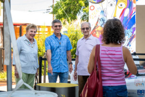 Four adults stand outdoors by colorful artwork, smiling and conversing. Two men and a woman face the camera; another woman with curly hair and a pink-striped shirt stands with her back to the camera.