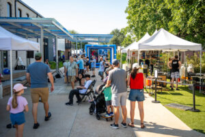 People walk through an outdoor market with white tents, vendors, and displays on a sunny day. Some visitors browse stalls, while others walk or sit, surrounded by trees and modern buildings.