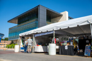 A white tent with art booths and people browsing is set up outside a modern building on a sunny day.