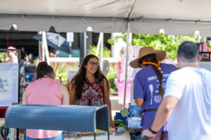 People shop at an outdoor art market under a tent. A smiling woman stands behind a table with art pieces, interacting with visitors. Bright sunny weather and colorful displays are visible in the background.