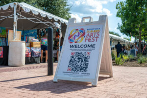 A sandwich board sign for the Round Rock Summer Arts Fest stands outdoors near vendor tents, displaying a QR code and festival information under a sunny sky.