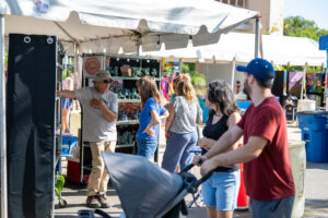People shop at an outdoor market under white tents. A vendor in sunglasses talks to shoppers browsing items on shelves. A man pushes a stroller while others walk by on a sunny day.