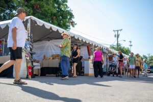 People browse vendor booths at an outdoor market under a white tent on a sunny day. Some wear hats and summer clothes, while others push strollers or carry bags. Trees and utility poles are visible in the background.