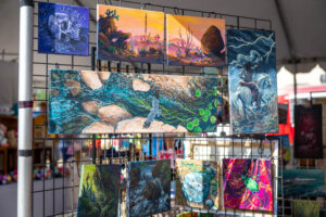 Colorful paintings are displayed on a metal grid at an outdoor art market, featuring nature scenes, animals, landscapes, and abstract art. The background shows market stalls under a tent.
