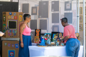 Two women stand at an outdoor art booth with colorful doodle portraits, framed art, and handmade crafts on display. A $6 Doodle Booth sign is visible on the left.