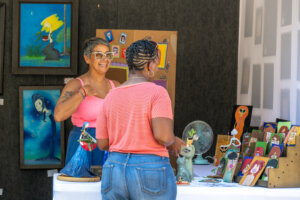 Two women talk at an art booth displaying colorful painted portraits, figurines, and framed artwork. One woman stands behind the table, smiling, while the other faces her, looking at the items.