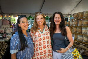 Three women smiling and posing together under a tent at an outdoor market, surrounded by tables with crafts and wooden items for sale.