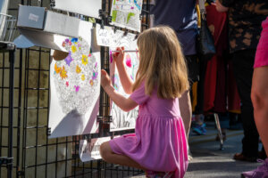 A young girl in a pink dress kneels on the ground, coloring a large, detailed illustration attached to a wire display at an outdoor event. Other people stand nearby.