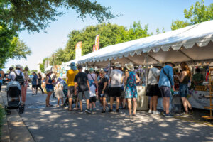 People line up at vendor booths under a white tent at an outdoor market on a sunny day. Some wear hats and summer clothes. Trees and signs are visible in the background.