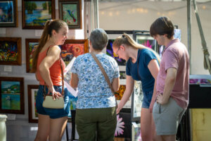 Four people look at paintings displayed at an outdoor art fair booth. Two young women, an older woman, and a young man are closely examining the colorful framed artwork under bright sunlight.