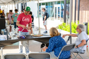 A man demonstrates sculpting to two seated people at an outdoor booth, with others walking and mingling in the background under a canopy.