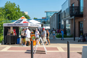 People gather around a white tent with Bring the Juice on it at an outdoor event on a sunny day, near modern brick buildings and greenery.