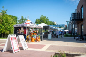 People gather under tents at an outdoor market on a sunny day. Signs and juice stand are in the foreground, while groups mingle near vendor stalls in the background. Trees and buildings line the area.