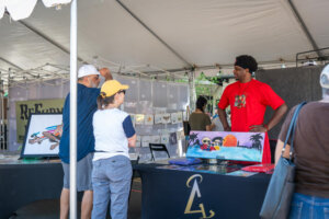 A man in a red shirt stands behind a table displaying colorful art at an outdoor market as two people look at the artwork and talk to him under a white tent.