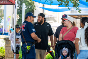 A firefighter in uniform smiles and talks with a group of people outdoors. A woman with a stroller, a baby, and others stand nearby at what appears to be a community event.