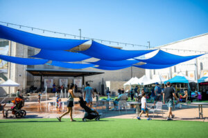 People stroll and relax under blue shade sails at an outdoor plaza with market tents, chairs, and a small splash pad on a sunny day.