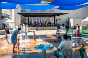 Children and adults play in a splash pad under blue sunshades, with people seated at tables nearby. A stage with banners and a few tents are visible in the background. Its a sunny day.