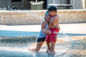 Two children play and laugh together in swimsuits at a splash pad, hugging and enjoying water spraying from the ground on a sunny day.